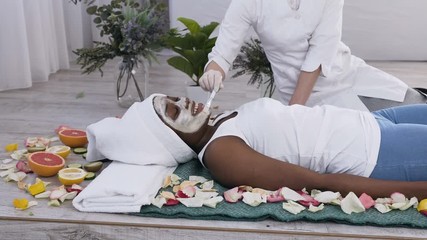 Pretty young african american woman with white towel on head lying relaxing on couch getting facial care by beautician in beauty salon, side view
