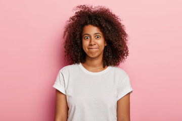 Photo of funny woman has curly thick hair, presses lips together, has happy face, wears white t shirt, isolated over pink background. Good looking young African American girl expresses happiness.
