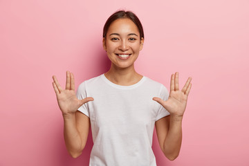 Young Asian female makes vulcan salute hand gesture, keeps arms raised and palms forward with extended thumbs, middle and ring fingers parted, greets you, says live long and prosper. Body language
