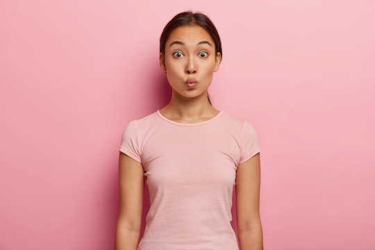 Lovely Asian girl with healthy skin, natural beauty, keeps lips folded for kiss, raises eyebrows, has dark hair combed in pony tail, wears casual outfit, models over pink wall. Human face expression