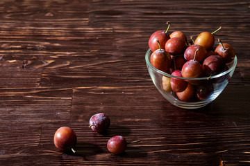 Ripe sweet plum fruits in glass bowl near with scattered plums on dark moody wood table background, hard light, copy space