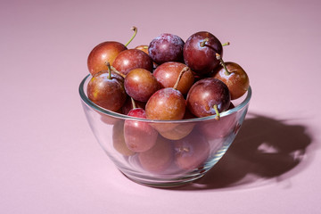 Ripe sweet plum fruits in glass bowl on pink background, hard light, copy space