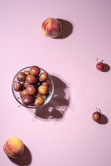 Ripe sweet plum fruits in glass bowl near with scattered plums and peaches on pink background, hard light 