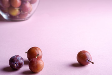 Scattered ripe sweet plum fruits close to glass bowl with plums on pink background, soft light, copy space, selective focus