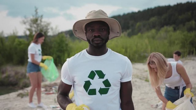 Outdoor portrait black young man eco activist collecting industrial waste from sandy beach cleaning up environment with a group of helpful activists. Fight pollution.