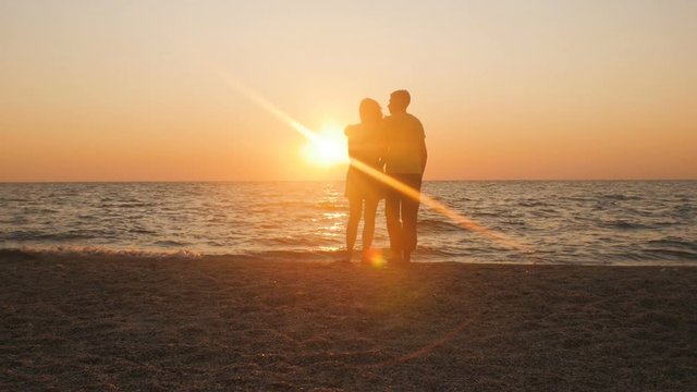 silhouette of a romantic young couple standing on the seashore, man hugging girl at sunrise on the beach, courtship on summer vacation and honeymoon