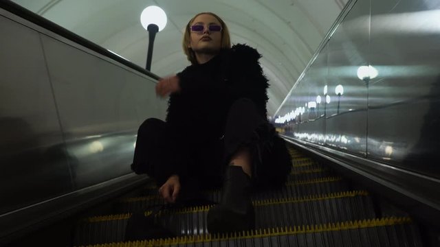 Cheeky young blonde with glasses. She is sitting on an escalator and posing for the camera.