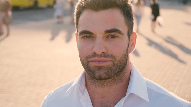 Portrait of young attractive man, wearing casual white shirt, man smiles and looks at the camera in city urban background at sunset. Close-up of Young handsome businessman of 30 years old outdoor.