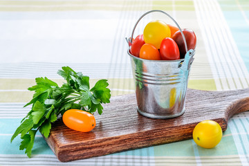 Ripe multicolored mini tomatoes in a small metal bucket and parsley over brown wooden cutting board on a tablecloth. Vegetables, vegetarian and healthy eating.