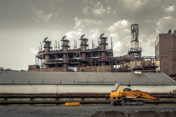 Yellow industrial machinery left empty in front of an abandoned factory with large smokestacks