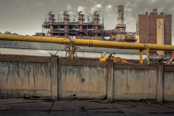 Large yellow pipe on top of a large concrete wall in front of giant smokestacks of an abandoned industrial factory