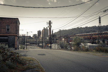 Looking down an empty street towards an abandoned industrial factory with overgrown vegetation