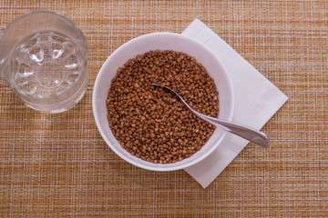 Bowl of raw buckwheat, cup of water and napkin on granny background, top view