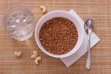 Bowl of raw buckwheat, cup of water, cashew nuts and napkin on granny background, top view