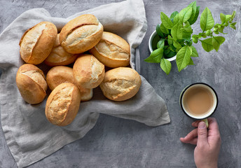Bread buns in basket on rustic wood, spring leaves and hand with cup of coffee