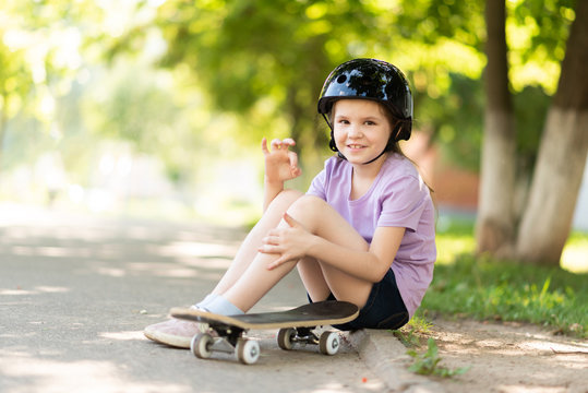 A little girl sits in a helmet and with a skateboard, and shows the sign okay.