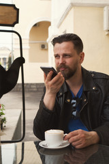 Stylish hipster freelancer drinking coffee on a summer terrace of a restaurant. A man is recording a voice message. Handsome guy with a fashionable haircut and neat beard