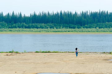  Girl tourist with a backpack , briskly goes one to a concrete cliff on the shore of the old pier at the Northern river vilyu with spruce tundra forests of Yakutia on the other side.