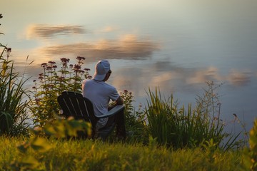 Man in the seat at bank of lake also looks afar on sunset, reflecting the shadow from the orange sky of the sunset. The film grain