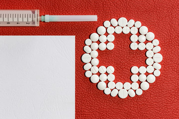 White medicinal pills, a syringe and a blank sheet of paper lie on the surface of red faux leather. Red Cross. Template for a recipe.