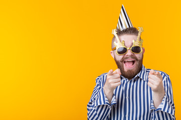 Joyful young hipster male in a paper cap and glasses is laughing happily on a yellow background with copy space. The concept of a mega party and holiday.