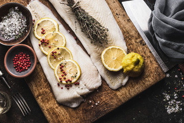 Close up of raw cod fillet with lemon slices and herbs on rustic wooden cutting board, top view....