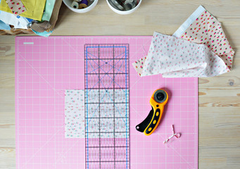 Work place: cotton fabrics, pink cutting mat, quilting ruler, rottary cutter, sewing supplies in white cups and wooden desk
