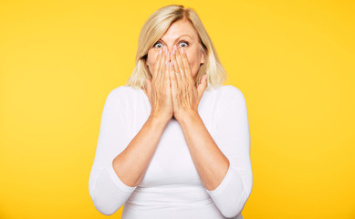 Don't talk! Blonde Senior woman covers her mouth with help of hands and looks on camera with shock in eyes. 