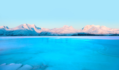 Cracks on the surface of the green ice next to red and orange houses (cabin) in winter -Beautiful landscape with Frozen lake - Tromso, Norway  M