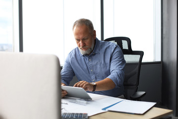 Mature businessman working using digital tablet while sitting in the modern office.