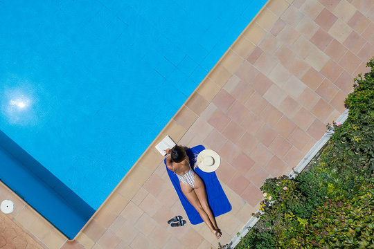 Mujer joven en ba&ntilde;ador tendida sobre una toalla de manera relajada en una piscina, vista cenital desde el aire
