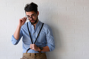 Portrait of handsome bearded hipster guy with glasses on standing indoors by the white brick wall