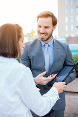 Happy young male and female business people meeting in front of an office building, talking and discussing. Woman is holding a tablet. Work anywhere concept.