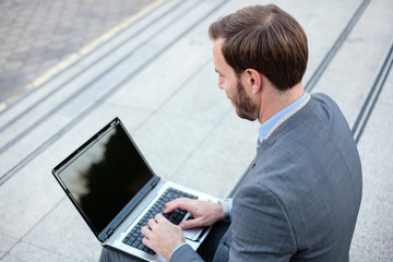 Handsome young businessman working on a laptop in front of an office building. High angle view over his shoulder. Work anywhere concept.