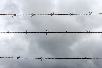 Barbed wire background. Three horizontal lines of barbed wire against a dramatic dark cloudy sky in monochrome colors. No access, trespassing or captivity symbol.
