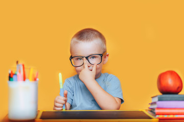 Happy blonde clever boy is sitting at a desk in glasses and smiling. Ready for school. Back to school. Apple, pens and books on desk
