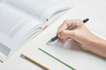 in school lesson. hand holds a pen near a squared notebook. next to it is a book and a pencil. white background