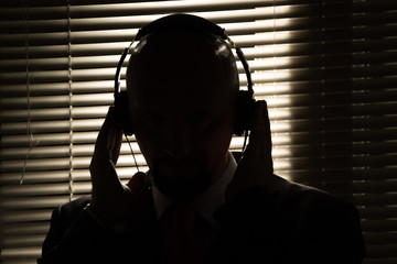 Bald man with headphones on the background of closed blinds, contour lighting, listening to music on old equipment - obrazy, fototapety, plakaty