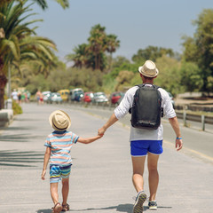 European dad and son in sunhats spending time together on summer vacations. They are holding hands. Back view.