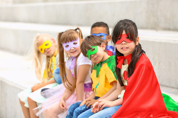 Cute little children dressed as superheroes sitting on stairs outdoors