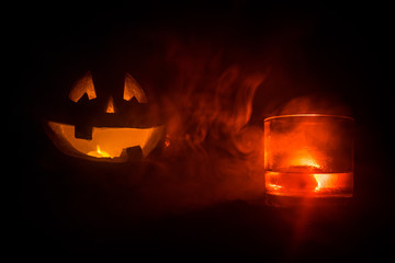 Halloween pumpkin with carved face and glass of whiskey with ice on a dark toned foggy background...