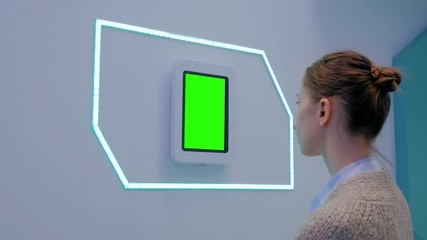 Woman looking at wall mounted white tablet with blank green display at exhibition or museum with futuristic sci-fi interior. Mock up, copyspace, template, chroma key, green screen, technology concept