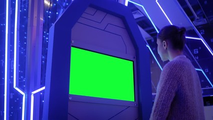 Woman looking at blank digital interactive green display kiosk at exhibition or museum with futuristic sci-fi interior. Mock up, copyspace, template, chroma key, green screen, technology concept