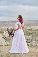 Fototapeta na wymiar Woman with beautiful bouquet of flowers in hands stands on mountain in rays of dawn sunset. Beautiful white long dress on the girl body. Perfect bride with pink hair. Turkey, Cappadocia