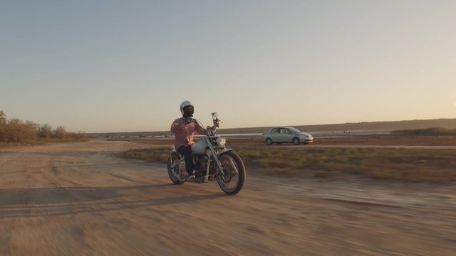 Young stylish motorcyclist driving his motorbike in the desert road during sunset 