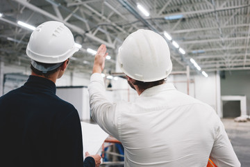 Two building engineers wearing safety hard hat discussing blueprint on construction site. Worker and inspector have meeting inside building under construction