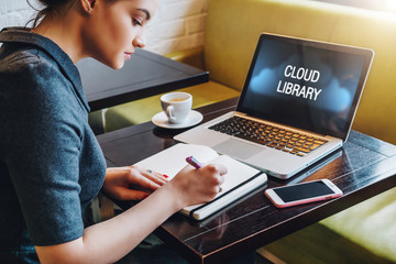 Woman sitting at table in cafe in front of laptop computer with inscription on monitor-cloud library and making notes in notebook.