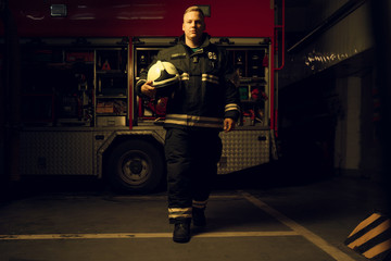 Full-length photo of young firefighter man with helmet in his hand looking into camera while