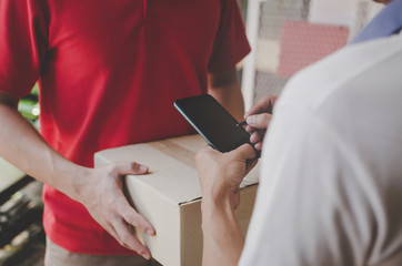 young man customer appending digital signature in mobile smart phone receiving parcel post box from courier with home delivery service man in red uniform at home, technology, express delivery concept
