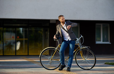 Fototapeta premium A young stylish bearded businessman in suit going to work by bike. Drinking coffee from a cup to go and talking on a mobile phone on the background of an office building. Takes a working break